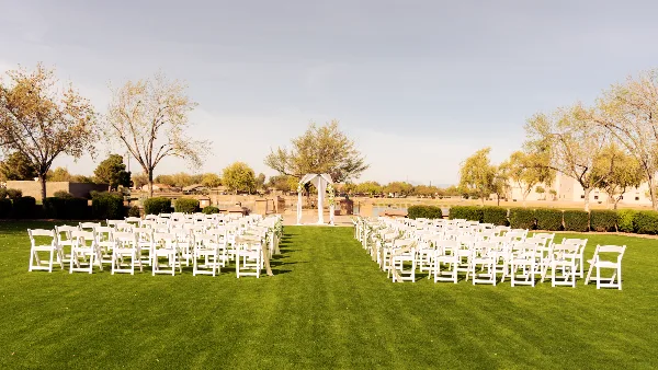 An outdoor wedding ceremony setup with rows of white chairs and a floral arch on a green lawn.