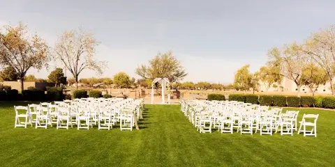 An outdoor wedding ceremony setup with rows of white chairs and a floral arch on a green lawn.