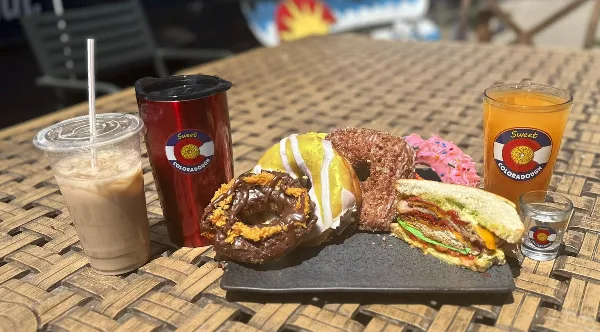 A table setting featuring assorted donuts, a sandwich, and branded coffee and juice cups from Sweet Coloradough.