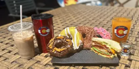 A table setting featuring assorted donuts, a sandwich, and branded coffee and juice cups from Sweet Coloradough.