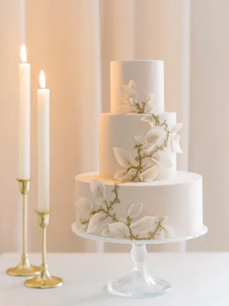 A three-tiered white wedding cake with sugar leaf decorations on a glass stand next to lit candles.