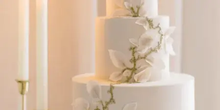 A three-tiered white wedding cake with sugar leaf decorations on a glass stand next to lit candles.
