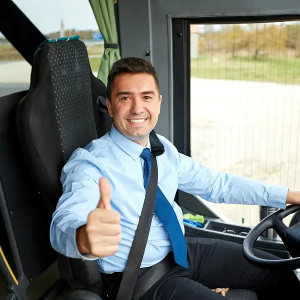 A smiling professional bus driver wearing a blue shirt and tie, seated in the driver's seat of a bus and giving a thumbs up.