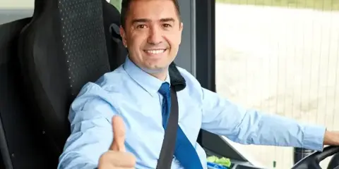 A smiling professional bus driver wearing a blue shirt and tie, seated in the driver's seat of a bus and giving a thumbs up.