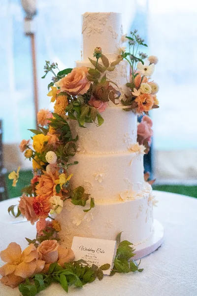 A four-tier white wedding cake decorated with cascading orange and yellow flowers and greenery, sitting on a table with a small card.