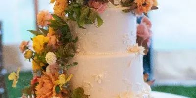 A four-tier white wedding cake decorated with cascading orange and yellow flowers and greenery, sitting on a table with a small card.