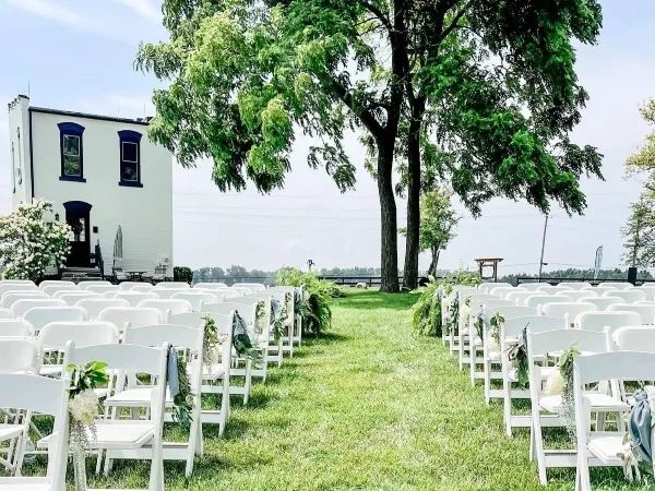Outdoor wedding ceremony setup with rows of white chairs decorated with greenery and flowers, a grass aisle, and a white historic building in the background.