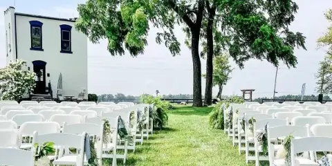 Outdoor wedding ceremony setup with rows of white chairs decorated with greenery and flowers, a grass aisle, and a white historic building in the background.