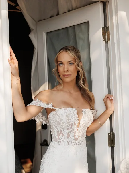 A bride in a white lace wedding dress standing in a doorway with professionally applied makeup and styled hair.