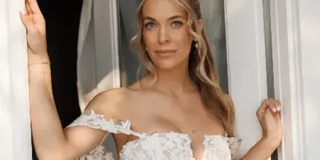 A bride in a white lace wedding dress standing in a doorway with professionally applied makeup and styled hair.