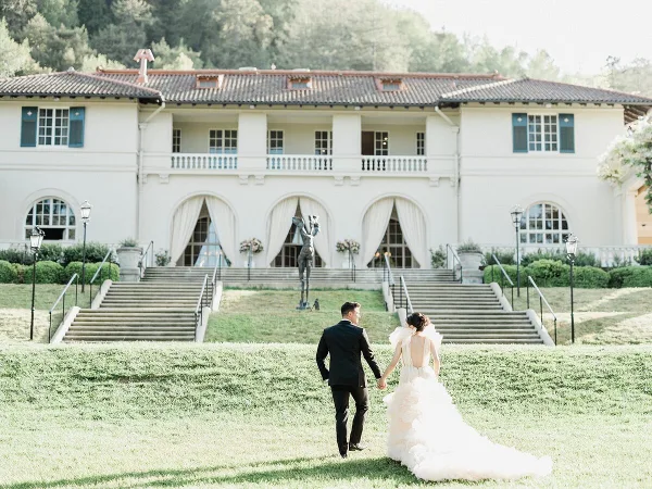 A bride and groom walking hand-in-hand across a lawn toward a large white wedding venue building.