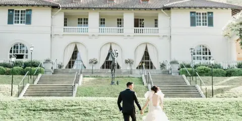 A bride and groom walking hand-in-hand across a lawn toward a large white wedding venue building.