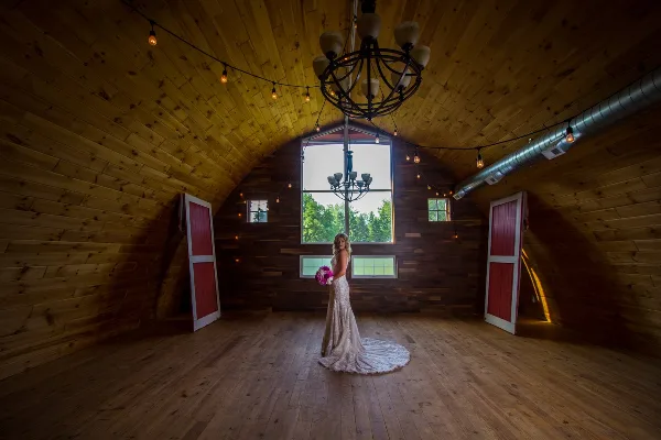 A bride in a white lace gown stands in a wooden barn venue with string lights and red doors.