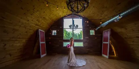 A bride in a white lace gown stands in a wooden barn venue with string lights and red doors.