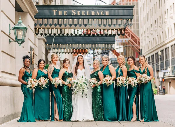 A bride in a white gown and veil stands with nine bridesmaids in emerald green dresses, all holding floral bouquets, in front of The Seelbach hotel.
