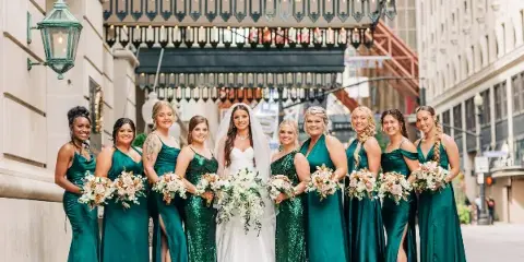 A bride in a white gown and veil stands with nine bridesmaids in emerald green dresses, all holding floral bouquets, in front of The Seelbach hotel.