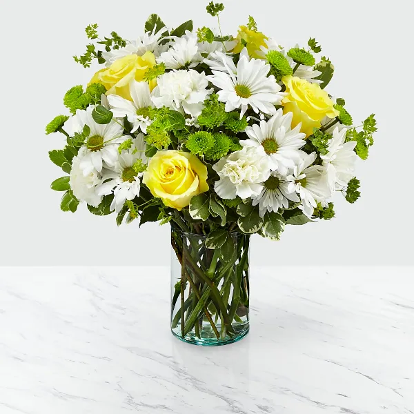 A mixed bouquet of white daisies, yellow roses, and green filler flowers arranged in a clear glass vase on a white marble surface.