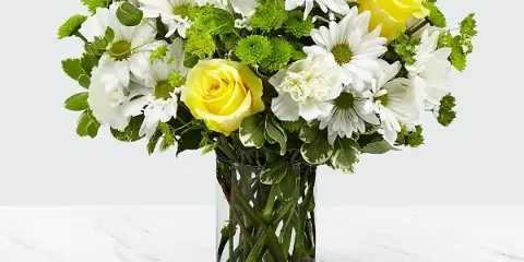 A mixed bouquet of white daisies, yellow roses, and green filler flowers arranged in a clear glass vase on a white marble surface.