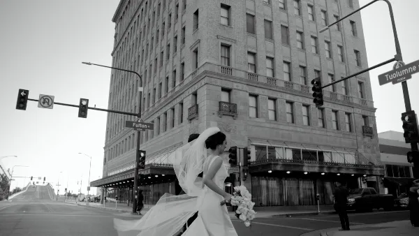 Black-and-white photo of a bride in a flowing gown and veil walking with her groom at a city street corner.