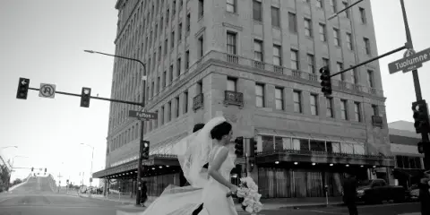 Black-and-white photo of a bride in a flowing gown and veil walking with her groom at a city street corner.