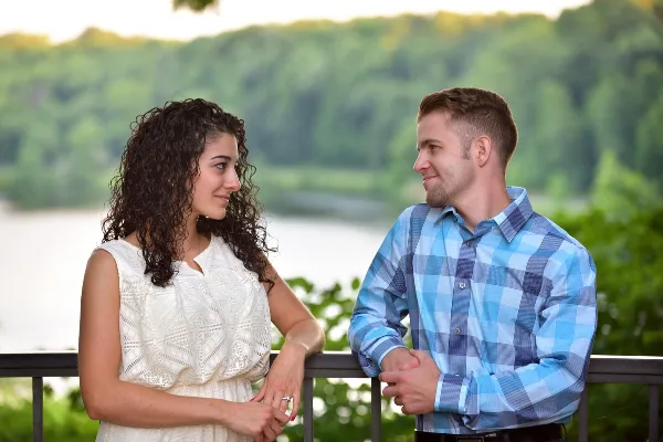 A professional outdoor engagement photograph of a couple standing on a balcony overlooking a river and green hills.