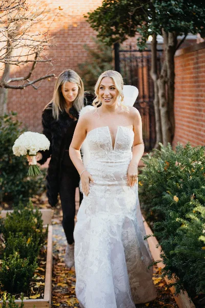 A bride in a white lace wedding dress walking through a garden path, accompanied by a woman in black holding a white rose bouquet.