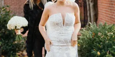 A bride in a white lace wedding dress walking through a garden path, accompanied by a woman in black holding a white rose bouquet.