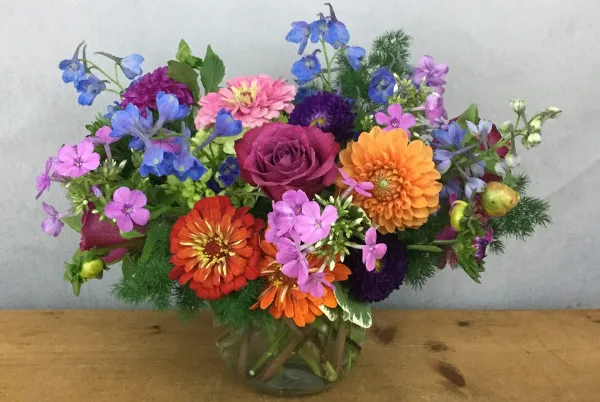 A colorful floral arrangement featuring roses, dahlias, and delphiniums in a glass vase.