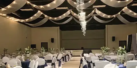 Interior view of a spacious event hall decorated with ceiling draping, a crystal chandelier, and round tables set for a wedding reception.