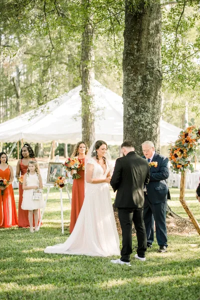 Outdoor wedding ceremony with a bride and groom exchanging vows under a white tent on a grassy farm.