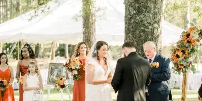 Outdoor wedding ceremony with a bride and groom exchanging vows under a white tent on a grassy farm.