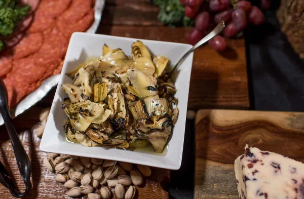 A white square bowl filled with marinated artichoke hearts sits on a wooden table next to a charcuterie board with salami, cheese, and grapes.