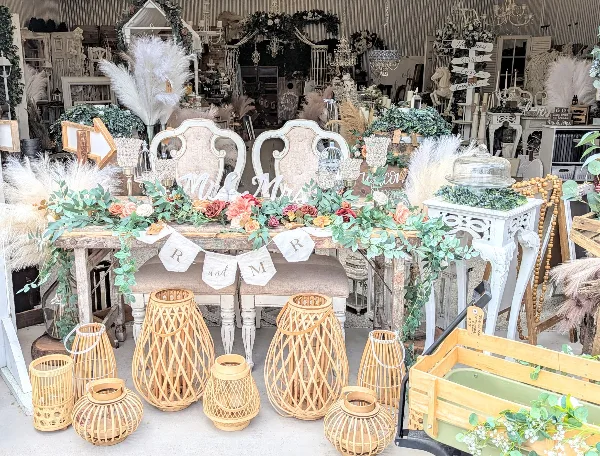 A wedding rental display featuring a rustic sweetheart table with floral garlands, white chairs, and bamboo lanterns.