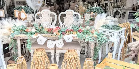 A wedding rental display featuring a rustic sweetheart table with floral garlands, white chairs, and bamboo lanterns.