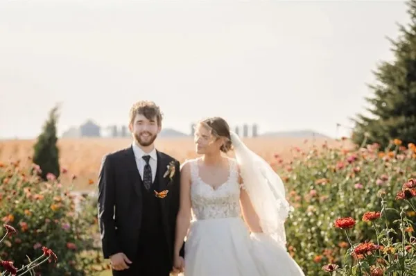 A bride and groom standing in a field of flowers with a barn visible in the background.
