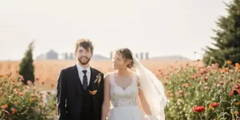 A bride and groom standing in a field of flowers with a barn visible in the background.