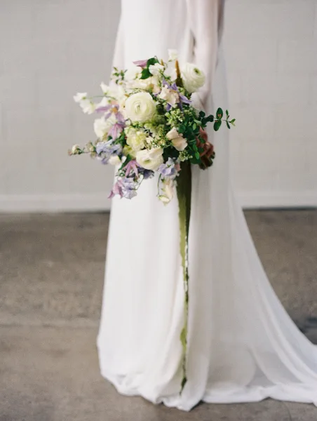 A close-up of a wedding bouquet held by a bride, featuring white ranunculus, roses, and purple delphiniums with greenery.