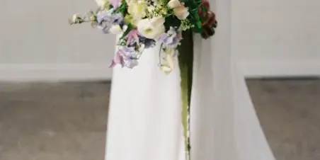 A close-up of a wedding bouquet held by a bride, featuring white ranunculus, roses, and purple delphiniums with greenery.