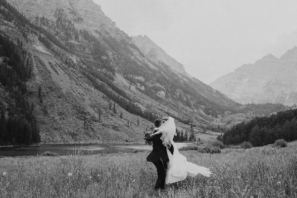 A black and white photograph of a bride and groom embracing in a grassy field with a mountain and lake in the background.