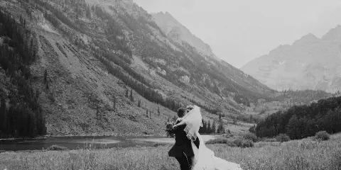 A black and white photograph of a bride and groom embracing in a grassy field with a mountain and lake in the background.