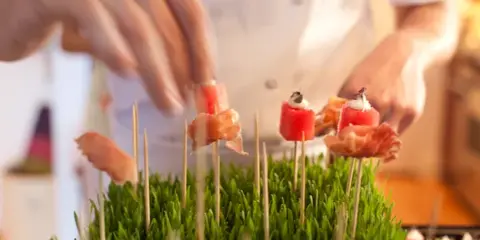 A chef's hands placing food skewers onto a bed of fresh green herbs.