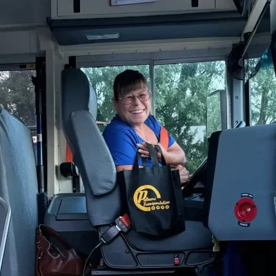 A smiling driver sitting in the driver's seat of a bus, holding a black Palmeri Transportation tote bag.