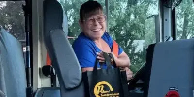 A smiling driver sitting in the driver's seat of a bus, holding a black Palmeri Transportation tote bag.