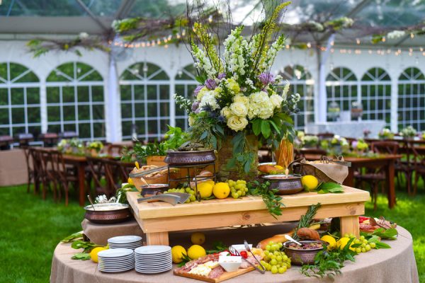 A catering table with a charcuterie board, fruit, and a floral centerpiece set up in a wedding venue.