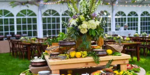 A catering table with a charcuterie board, fruit, and a floral centerpiece set up in a wedding venue.