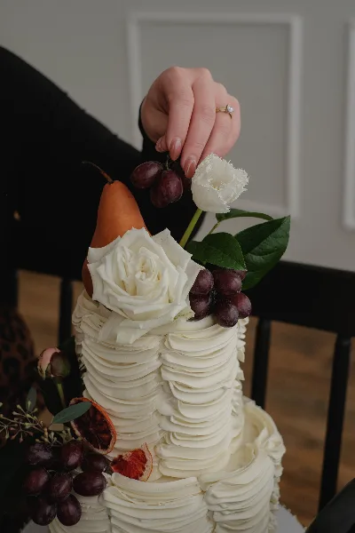 A hand placing a white flower and grapes onto a tall, white buttercream wedding cake.