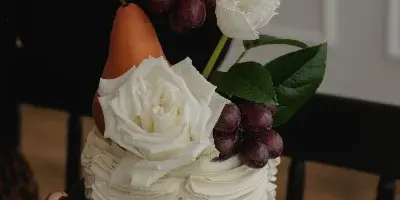 A hand placing a white flower and grapes onto a tall, white buttercream wedding cake.