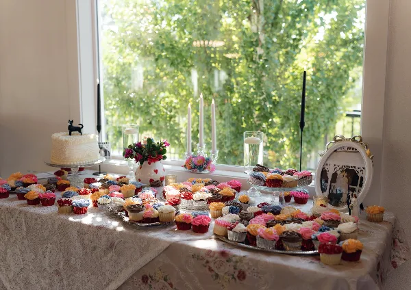 A wedding dessert table with a white cake, colorful cupcakes, and floral centerpieces arranged on a lace tablecloth near a window.
