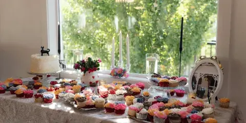 A wedding dessert table with a white cake, colorful cupcakes, and floral centerpieces arranged on a lace tablecloth near a window.