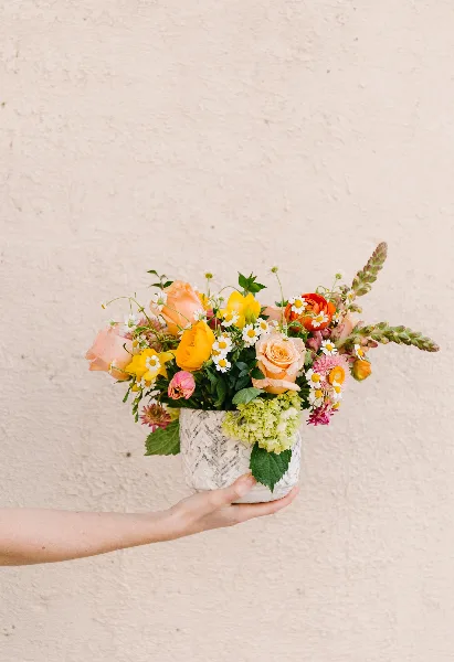 A hand holding a vase filled with a colorful bouquet of fresh flowers including roses, tulips, and daisies against a textured white wall.